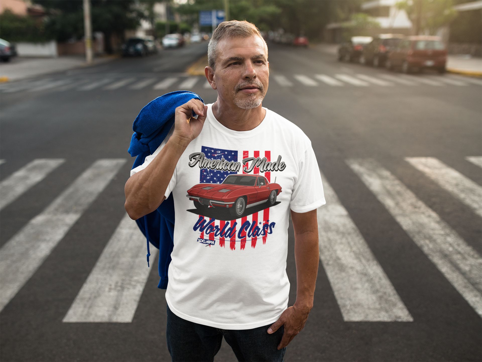Image of A man standing in the middle of the road, with a jacket over his shoulder. He is wearing a white tee shirt with the image of a Mustang muscle car. The shirt has text that reads 