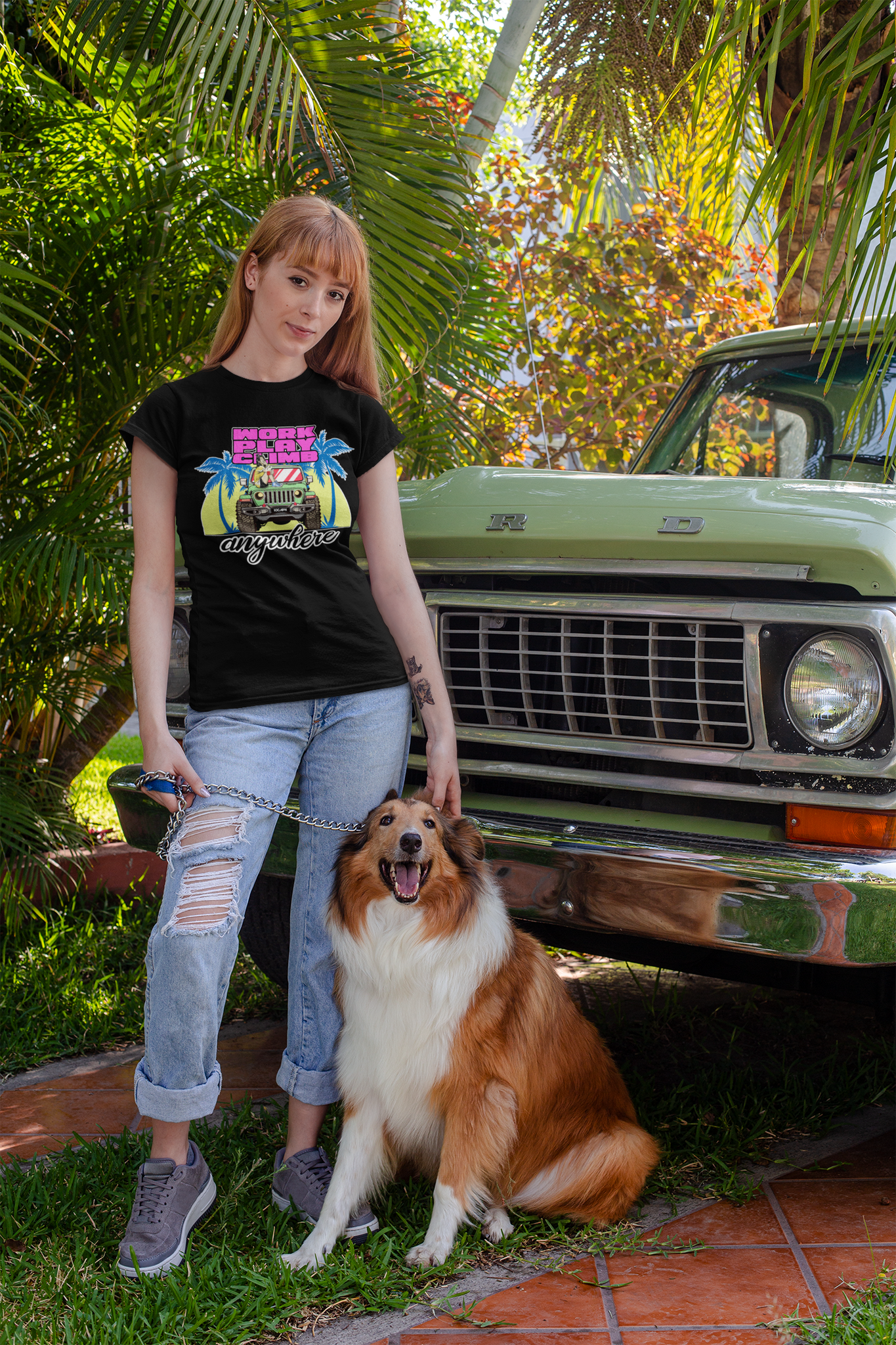 A woman standing with her dog in front of a truck. She is wearing a black tee shirt with the image of a Jeep Wrangler, and text reading "Work, Play, Climb Anywhere"