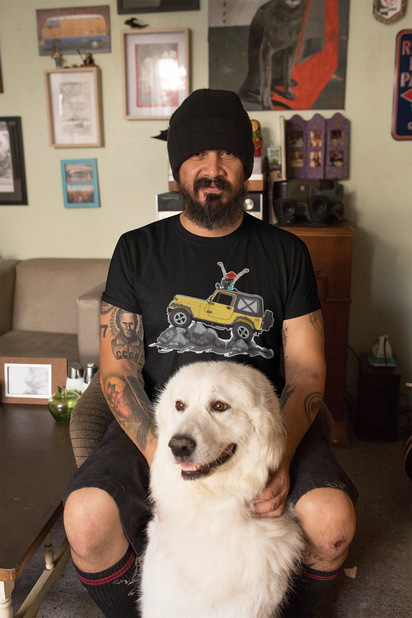 Image of A man sitting in a living room with a white dog. The man is wearing a black tee shirt with the image of a Jeep Gladiator climbing a mountain.