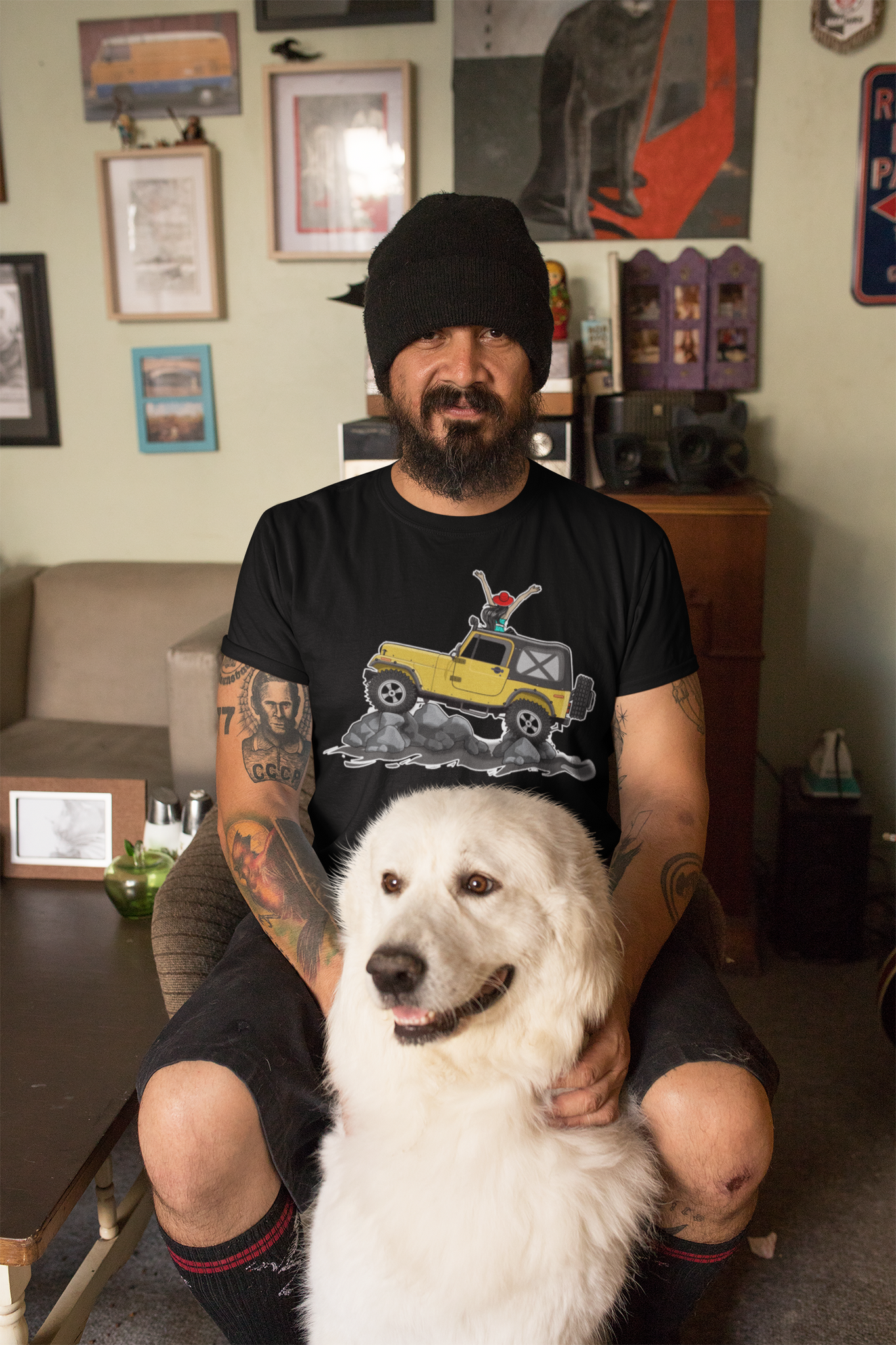 A man sitting in a living room with a white dog. The man is wearing a black tee shirt with the image of a Jeep Gladiator climbing a mountain.