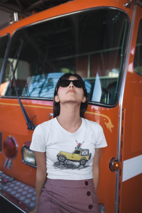 A woman in sunglasses standing in front of an orange van. She is wearing a white tee shirt with the image of a Jeep Gladiator climbing a mountain.