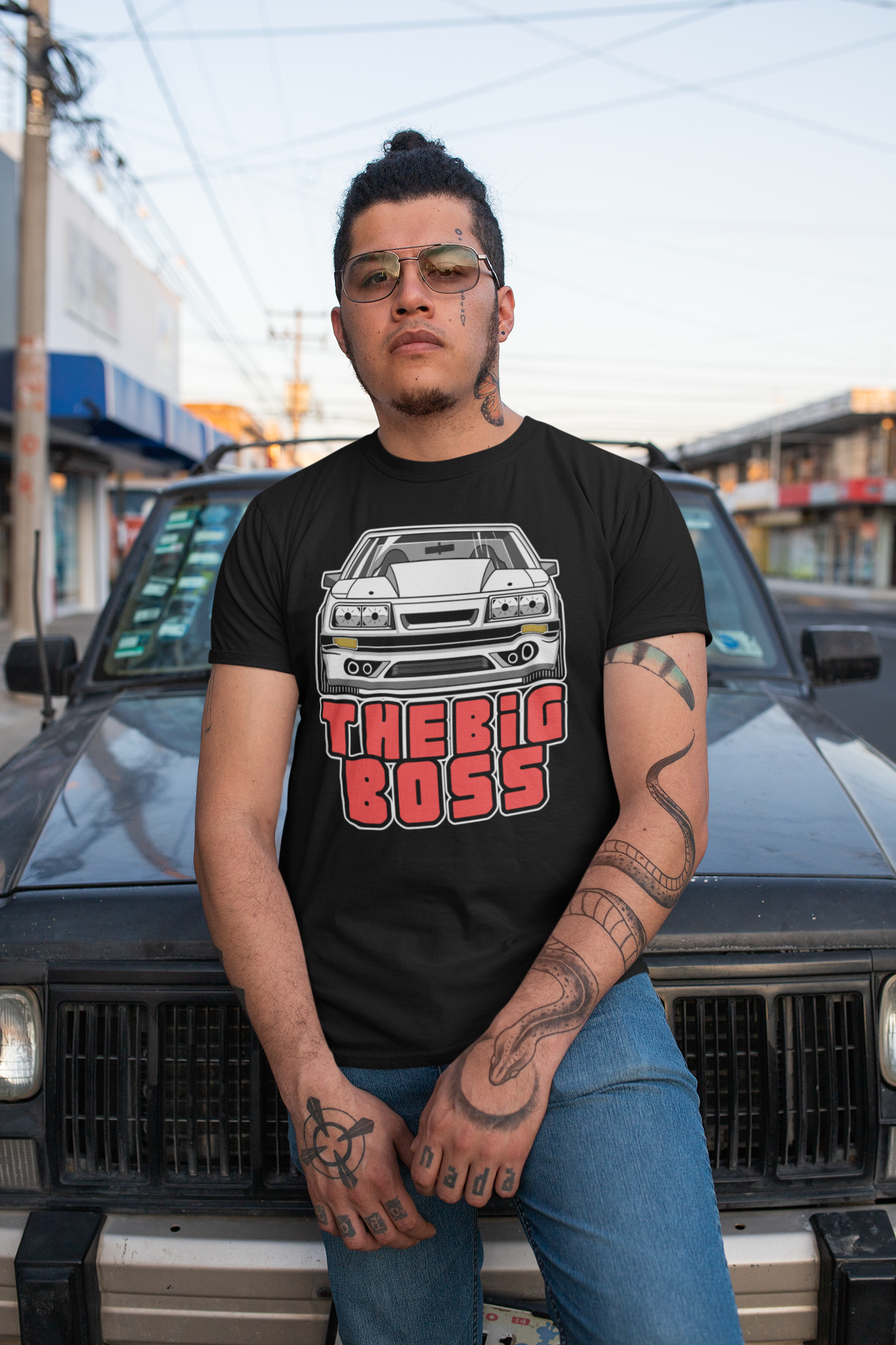 Image of A man leaning against a car, while wearing a black tee shirt with an image of a Ford Mustand Foxbody, and text reading 