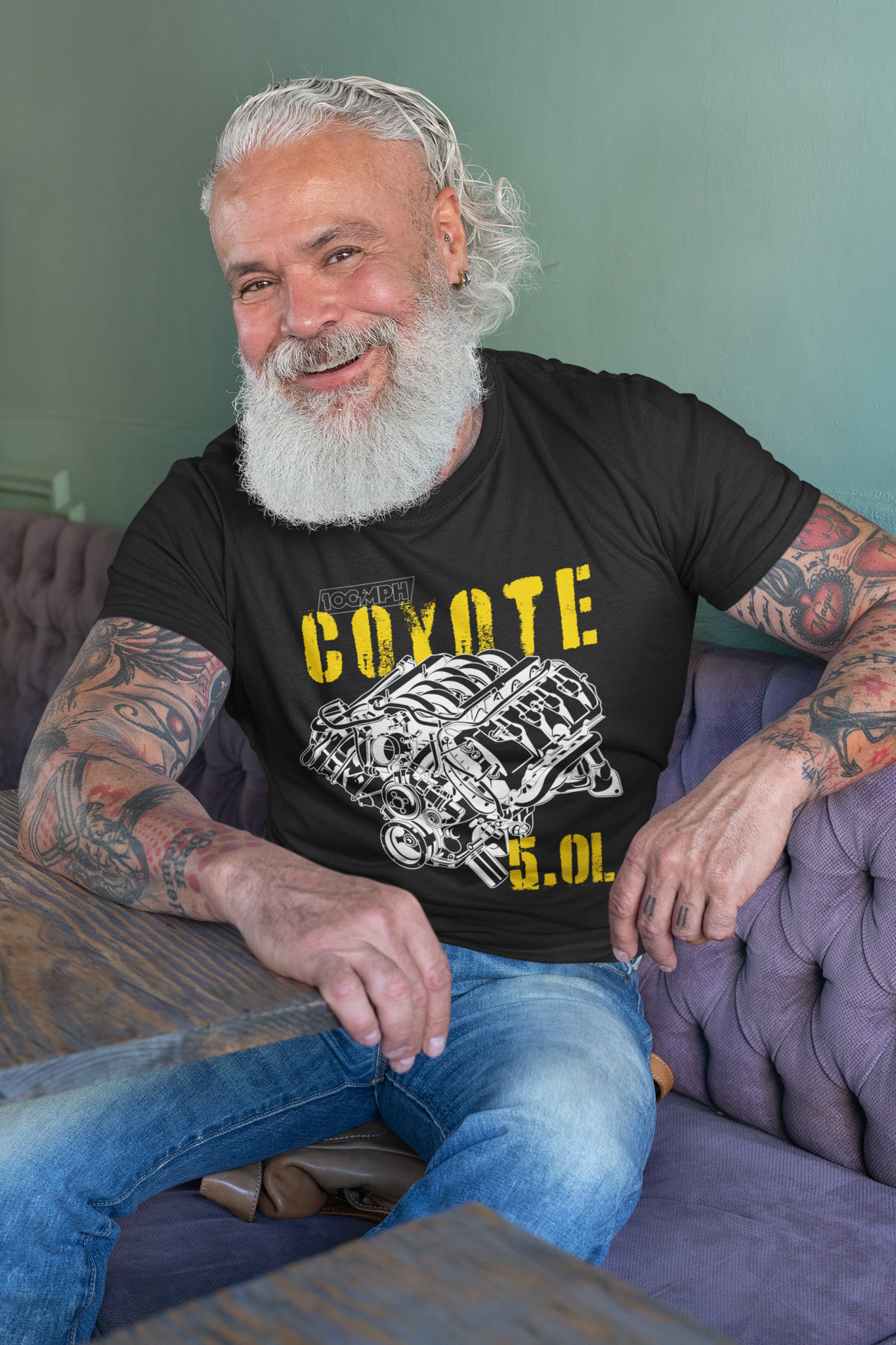 A man sitting in a restaurant leaning against a table. He's wearing a black tee shirt with an image of a Coyote engine. The tee shirt has text that reads "Coyote 5.0L". 